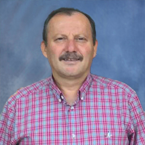 Portrait of a middle-aged man with a mustache, wearing a pink plaid shirt, standing against a blue studio backdrop.