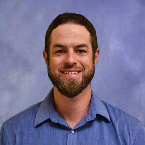 Portrait of a smiling man with a dark beard and short hair, wearing a blue collared shirt.