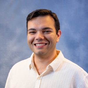 Young man smiling at the camera against a blue studio backdrop, wearing a light textured shirt.