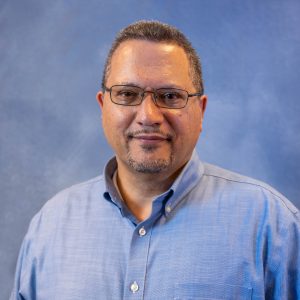 Portrait of a man wearing glasses and a light blue button-down shirt against a blue backdrop, looking at the camera.