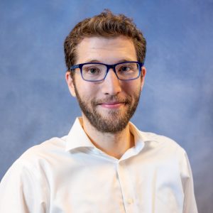 Portrait of a man with brown hair and beard wearing blue-framed glasses and a white shirt against a blue backdrop.