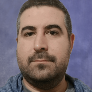 Close-up portrait of a man with short dark hair and beard, wearing a blue hoodie, against a blue studio backdrop.