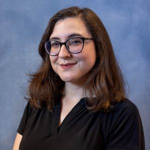 Portrait of a smiling woman with dark hair and glasses, wearing a black blouse, against a blue studio backdrop.