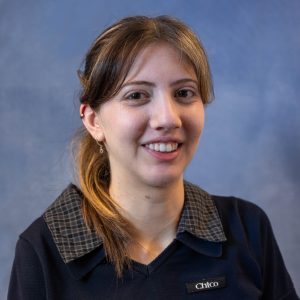 Smiling woman wearing a navy sweater with a plaid collar; name tag says Chico, against a blue backdrop.