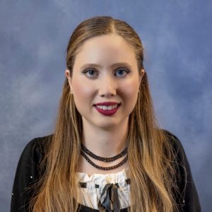 Portrait of a young woman with long light-brown hair, wearing a black top with a white ruffled collar, against a blue studio background.